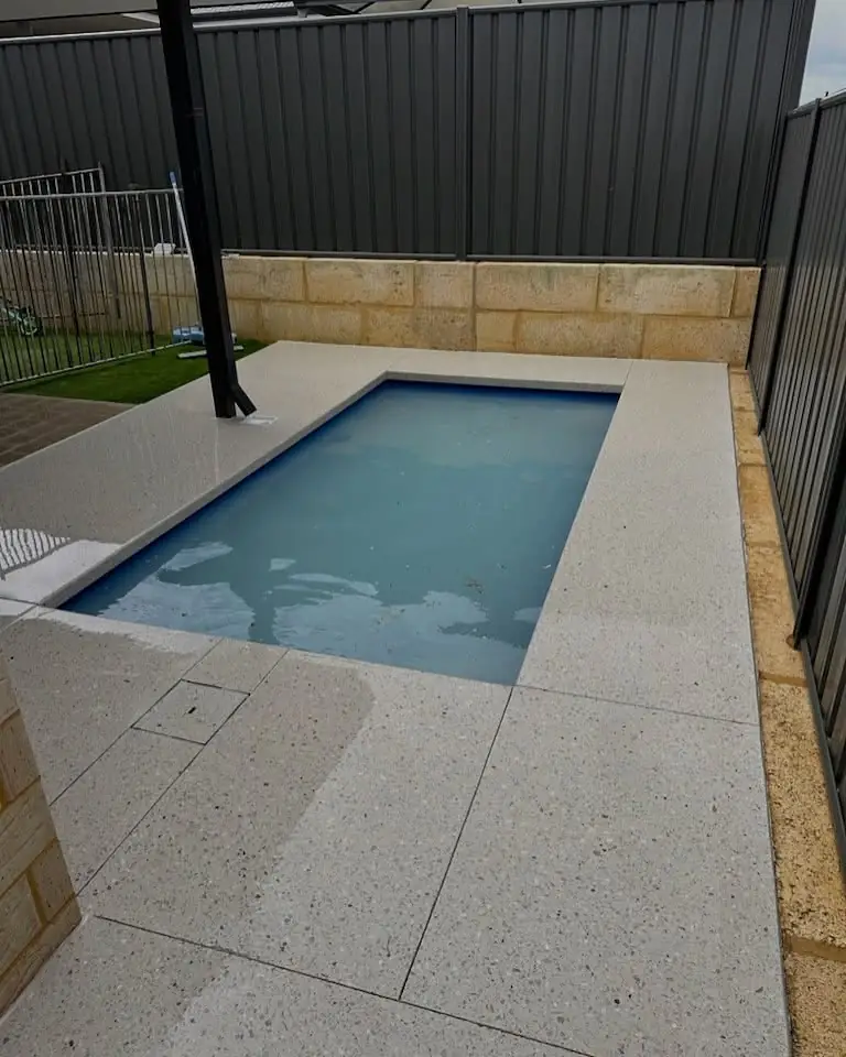 Honed white aggregate pool surround against a limestone wall, with a lawn strip and pool fence to the side