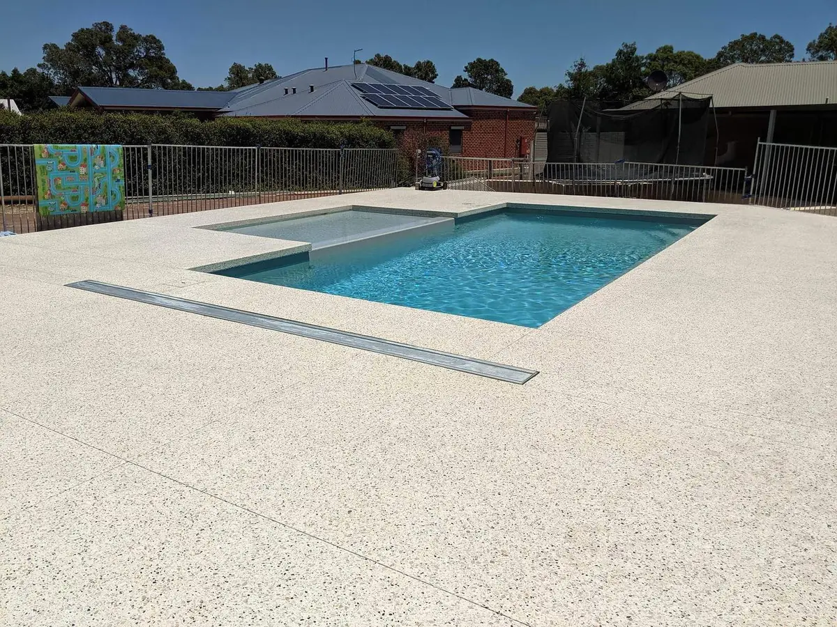 White exposed aggregate pool surround with a step and a bench built into the pool, finished and ready to swim