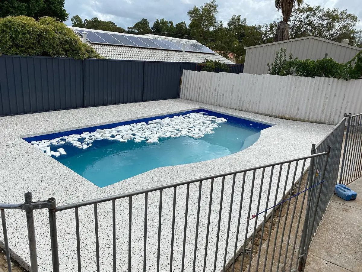 Pool surround in white exposed aggregate with a pool fence in the foreground and a plunge pool full of water toys